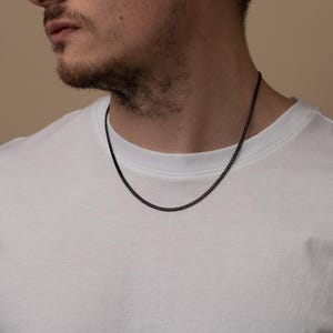 May include: A black curb chain necklace. The necklace is worn over a white t-shirt. The chain is made of a dark metal. The chain's links are closely connected.