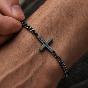 May include: A dark gunmetal gray chain bracelet featuring a central cross charm. The cross is engraved with the Roman numerals XII.IV.MCMXCVI. The bracelet consists of small, interlocking metal links that form a continuous band around the wrist.