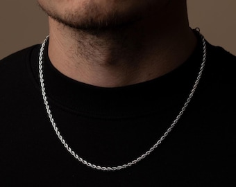 May include: A silver rope chain necklace. The necklace is made of a twisted, rope-like design. The chain is worn around the neck, against a black shirt. The background is a neutral color.