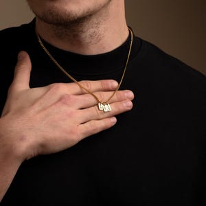May include: A gold-colored chain necklace with three small rectangular pendants. Each pendant has engraved text. The necklace is worn against a black shirt. The person's hand is resting on the necklace.