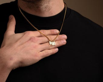 May include: A gold-colored chain necklace with three small rectangular pendants. Each pendant has engraved text. The necklace is worn against a black shirt. The person's hand is resting on the necklace.