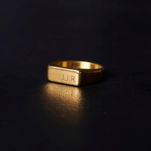 Peut inclure: Une bague chevalière dorée avec une face rectangulaire. Les initiales "J.J.R" sont gravées sur la face de la bague. La bague est présentée sur un fond sombre, soulignant sa surface polie et son design. Bijou classique.