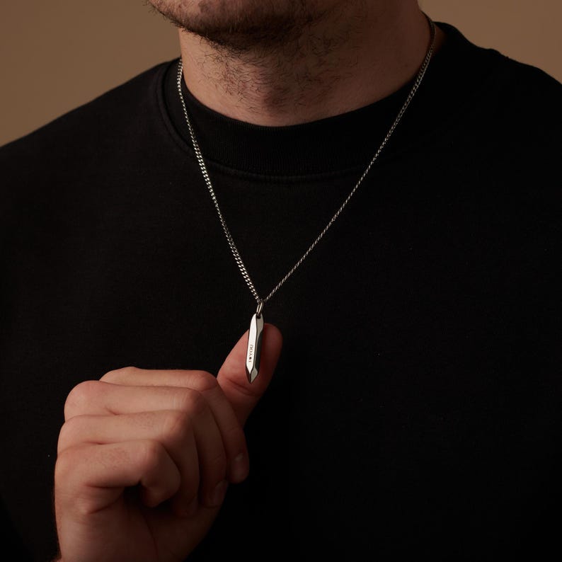 May include: A silver-colored pendant necklace with a geometric design. The pendant is a long, slender shape with a textured surface. The necklace chain is a simple curb style. The person is wearing a black shirt.