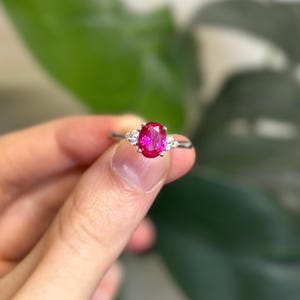 以下が含まれることがあります： 鮮やかなオーバルカットのルビーの宝石をあしらったシルバーリング。両側に2つの小さな白い宝石が配置されています。ぼやけた緑色の背景にリングが配置されています。