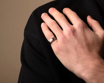 May include: A silver ring with a textured surface. The ring is worn on a finger. The ring is a band style, with a hammered or textured finish. The ring is made of a shiny metal.