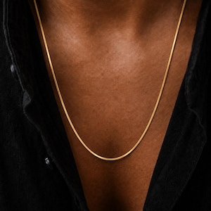 Puede incluir: Un collar de cadena tipo serpiente, plano y de tono dorado, que descansa sobre la piel. El collar presenta una superficie metálica lisa y continua con un acabado reflectante, usado con una camisa oscura de cuello abotonado a los lados.