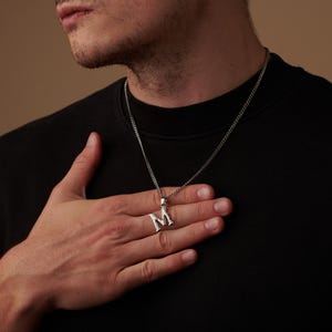 Puede incluir: Collar con colgante inicial plateado con la letra "M". El colgante cuelga de una cadena fina. El collar se lleva sobre una camisa negra.