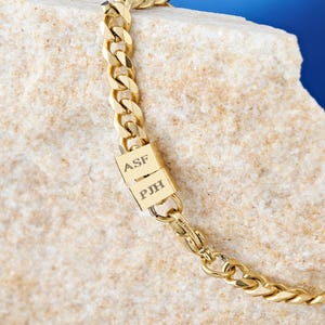 Può includere: Bracciale a catena color oro con design a maglie a catena. Il bracciale presenta due chiusure rettangolari con le iniziali "ASF" e "PJH" incise. Il bracciale è esposto su una superficie chiara e strutturata.