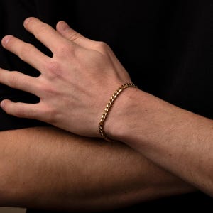 May include: A gold-colored curb chain bracelet. The bracelet is made of interlocking links and is worn on a person's wrist. The background is black.
