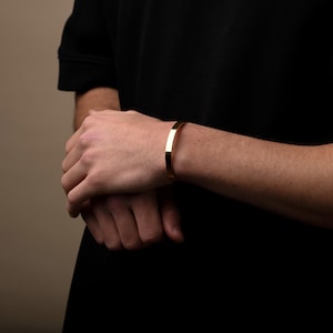 May include: A gold-coloured, rectangular bracelet. The bracelet is worn on a person's wrist. The background is a solid, neutral colour. The bracelet is a simple, elegant design.