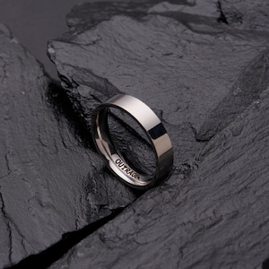 Puede incluir: Un anillo plateado con una superficie plana y pulida. La palabra "OUTRAGE" está grabada en el interior del anillo. El anillo se muestra sobre un fondo oscuro y texturizado. Diseño sencillo y moderno.