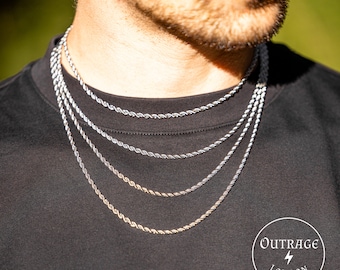 May include: A close-up of a man wearing a black t-shirt with four different silver rope chains layered around his neck. The chains are all different thicknesses and have a shiny finish. The image is cropped at the shoulders and the man's face is visible. The text "OUTRAGE LONDON" is visible in the bottom right corner of the image.