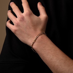 May include: A gold-colored Figaro chain bracelet. The bracelet is made of interlocking oval and elongated links. The bracelet is worn on a wrist against a black background.