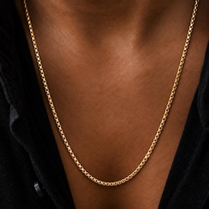 Peut inclure: Un collier chaîne de type boîte en ton doré composé d'une série de maillons carrés interconnectés formant un brin lisse et continu. La chaîne repose sur la peau, mettant en valeur une finition métallique polie et un motif géométrique uniforme.
