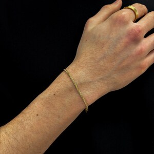 Pode incluir: Uma pulseira de corrente de corda dourada. A pulseira é usada em um pulso e o fundo é preto. Um anel de ouro está na mão.