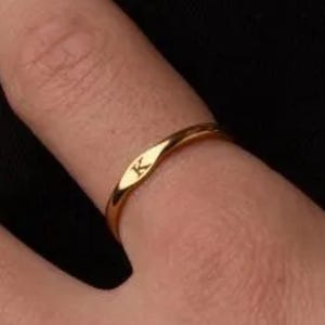 May include: A gold ring with a thin band. The ring has a small, elongated section with the letter "K" engraved. The ring is on a finger, set against a dark backdrop.