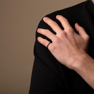 以下が含まれることがあります： 黒い指輪が指に着用されています。リングはシンプルなバンドデザインです。その人は黒いシャツを着ています。背景は無地のタン色です。