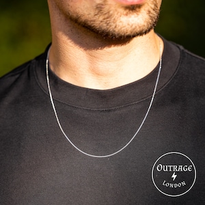 Könnte beinhalten: Eine silberne Kettenkette, die von einer Person mit einem schwarzen T-Shirt getragen wird. Die Kette ist dünn und zart und ein schlichtes und elegantes Schmuckstück.  Outrage London ist auf dem T-Shirt aufgedruckt.