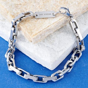 May include: A silver-toned bracelet with a chain link design. The bracelet features a rectangular tag with the inscription "MI VIDA". The bracelet is displayed on a white and beige surface against a blue background.