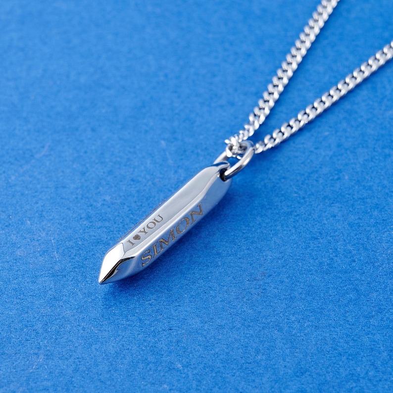May include: A silver-colored pendant necklace with a pointed, bullet-shaped charm. The charm is engraved with the name "SIMON" and the words "I YOU". The necklace chain is a delicate, linked design, set against a blue background.