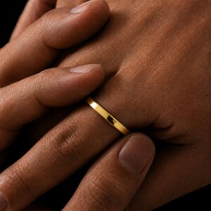May include: A simple, polished gold band ring with a smooth, flat surface and a thin profile. The ring features a continuous, unbroken circular design with a reflective metallic finish that catches the light, showcasing a clean and understated aesthetic.