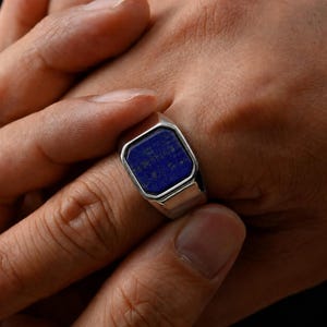 May include: A silver-toned signet ring featuring a rectangular, deep blue lapis lazuli gemstone inlay. The metal band has a polished, smooth finish with a flat, beveled setting that secures the dark blue stone in the center of the ring face.