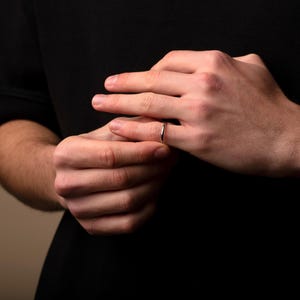 May include: A silver ring is worn on a finger. The ring is thin and has a polished finish. The hands are in focus against a dark background. The ring is a simple band, likely for everyday wear.
