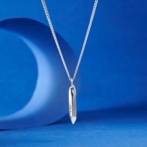 May include: A silver-colored necklace with a pendant. The pendant is a long, slender shape with the word "JOHN" engraved on it. The necklace chain is a delicate curb link style. The background is a gradient of blue.