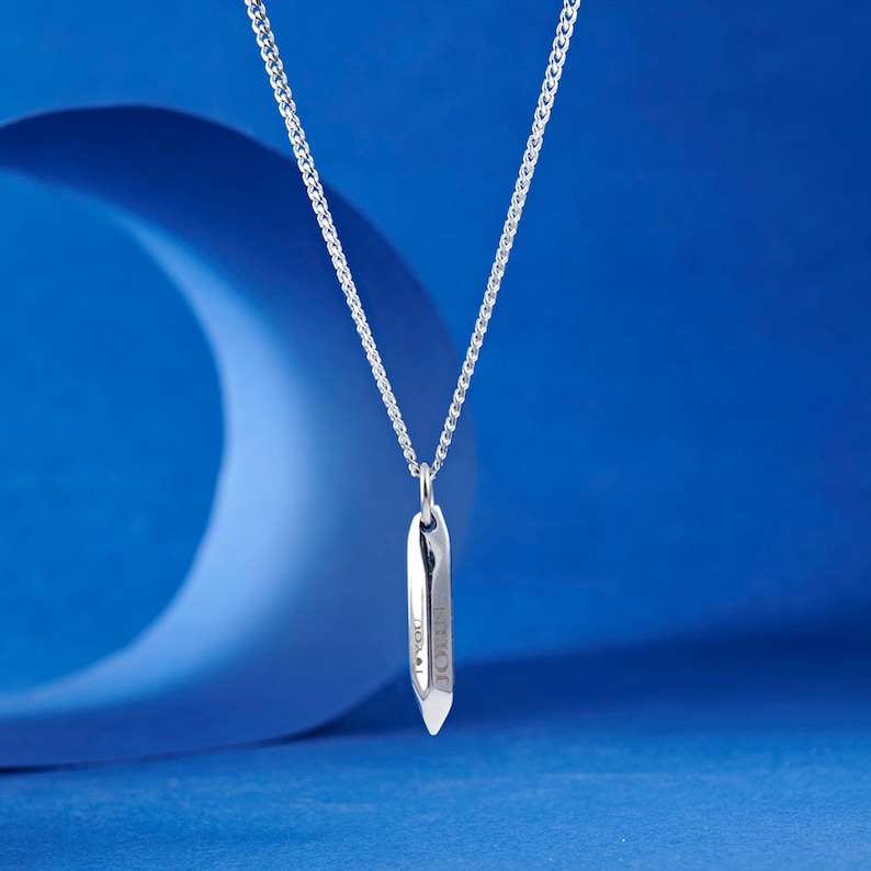 May include: A silver-colored necklace with a pendant. The pendant is a long, slender shape with the word "JOHN" engraved on it. The necklace chain is a delicate curb link style. The background is a gradient of blue.