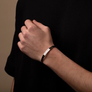 May include: A silver-colored cuff bracelet with the inscription "V.X.MCMCI" is worn on a wrist. The bracelet is a simple, minimalist design, reflecting a modern aesthetic. The background is a dark, neutral tone, highlighting the jewelry.