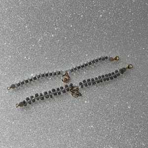 May include: Two gray beaded bracelets with gold charms. One charm is a crescent moon with a star, the other is a stylized leaf.