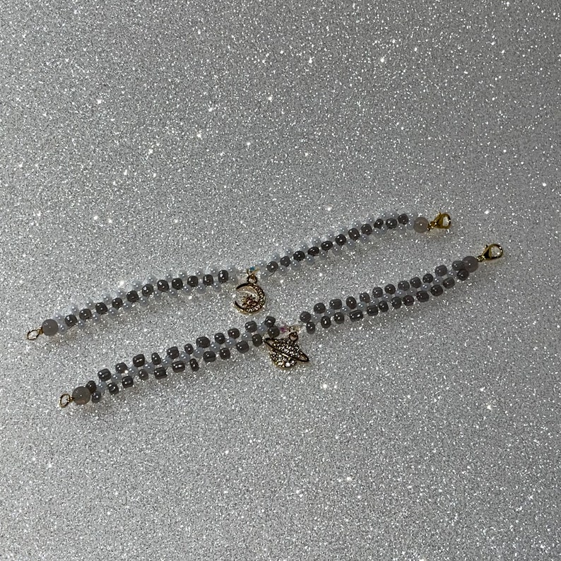 May include: Two gray beaded bracelets with gold charms. One charm is a crescent moon with a star, the other is a stylized leaf.
