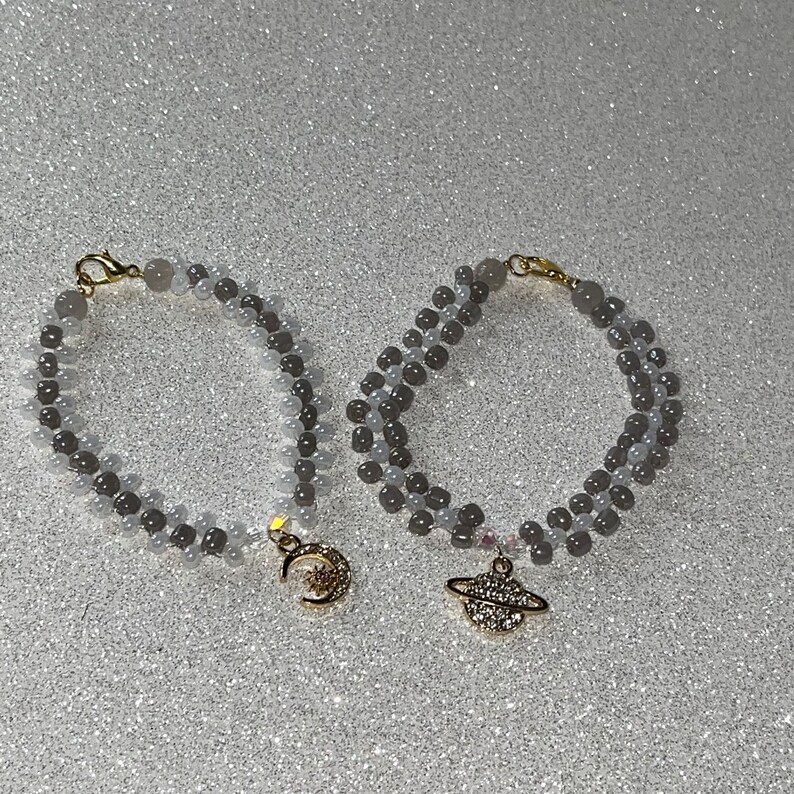 May include: Two gray beaded bracelets with gold charms. One charm is a crescent moon with a star inside. The other charm is a ringed planet with a star inside.