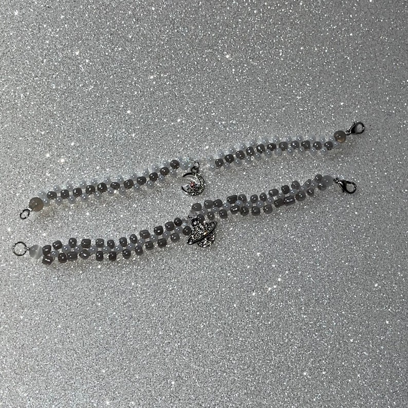 May include: Two silver beaded bracelets with celestial charms. One charm is a crescent moon and the other is a planet with a ring.