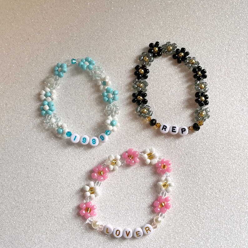 Pu&ograve; includere: Tre braccialetti di perline con ciondoli a forma di fiore e perline con lettere che compongono le parole "1989", "REP" e "LOVER". I braccialetti sono realizzati con perline bianche, blu, rosa, nere e dorate.