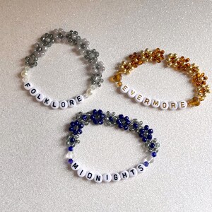 Pu&ograve; includere: Tre braccialetti di perline con perline di lettere bianche che compongono le parole "folklore", "evermore" e "midnights". Le perline sono grigie, dorate e blu.