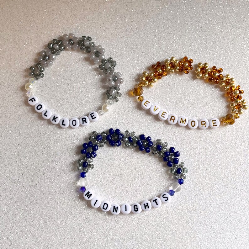 Pu&ograve; includere: Tre braccialetti di perline con perline di lettere bianche che compongono le parole "folklore", "evermore" e "midnights". Le perline sono grigie, dorate e blu.