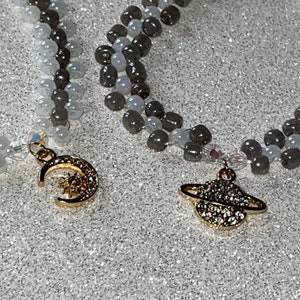 May include: Two necklaces with gray and white beads. One necklace has a gold crescent moon charm with a sun inside. The other necklace has a gold charm of a planet with a ring around it.