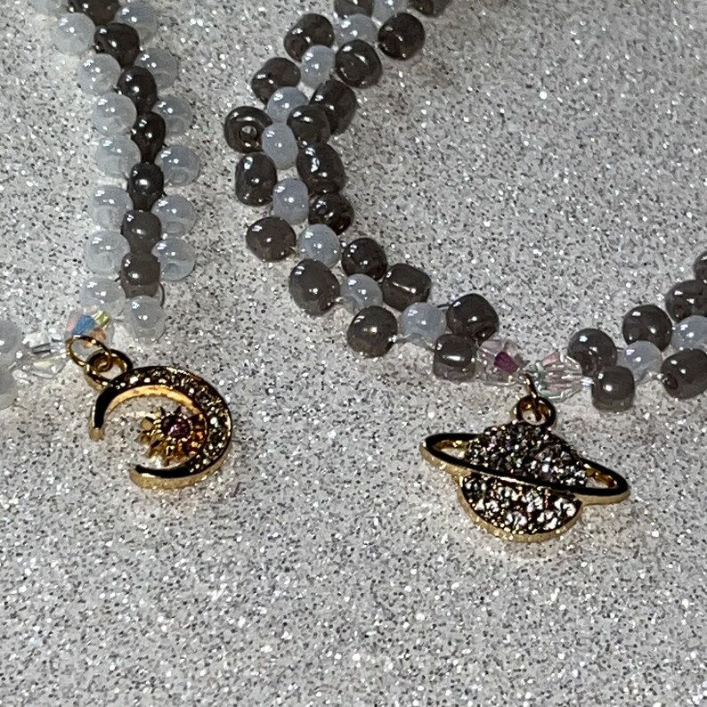 May include: Two necklaces with gray and white beads. One necklace has a gold crescent moon charm with a sun inside. The other necklace has a gold charm of a planet with a ring around it.