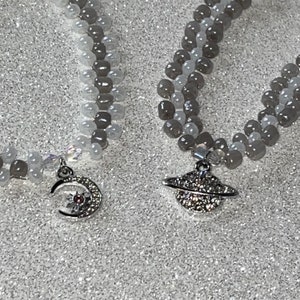 May include: Two beaded bracelets with silver charms. One charm is a crescent moon with a star and a pink gem. The other charm is a silver planet with a ring and rhinestones.