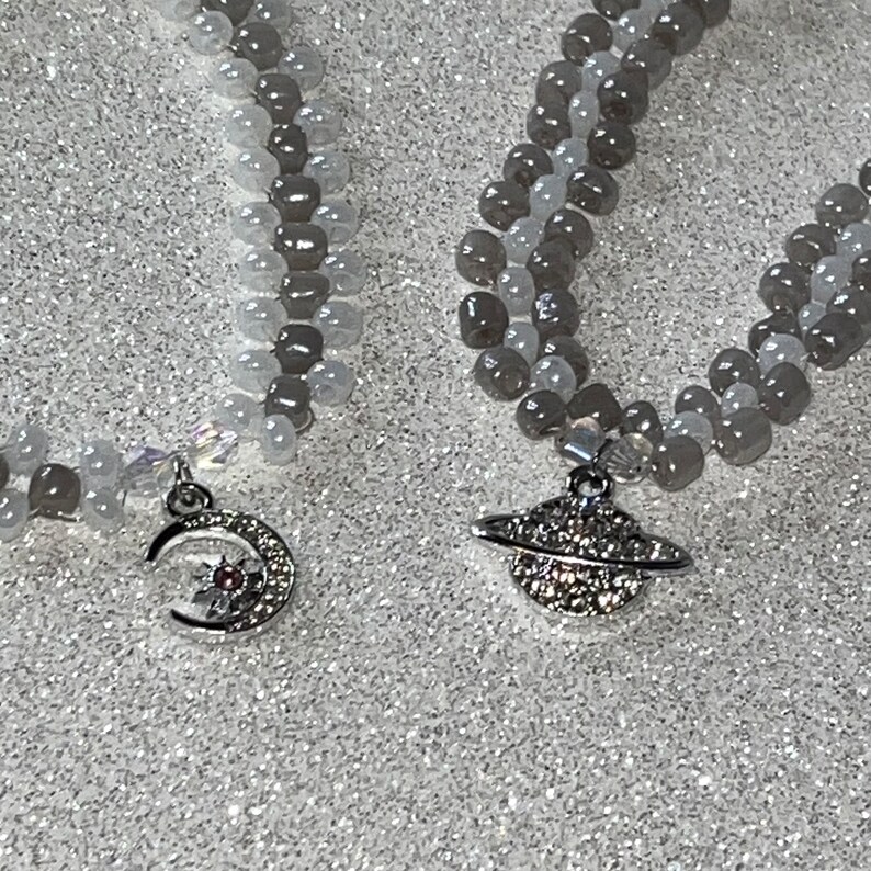 May include: Two beaded bracelets with silver charms. One charm is a crescent moon with a star and a pink gem. The other charm is a silver planet with a ring and rhinestones.