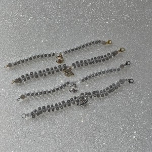 May include: Five gray beaded bracelets with gold charms. The charms include a crescent moon, a star, and a planet. The bracelets are layered on a white sparkly background.