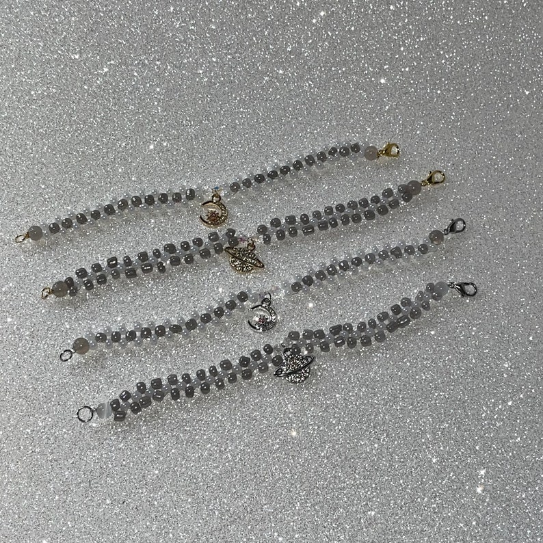 May include: Five gray beaded bracelets with gold charms. The charms include a crescent moon, a star, and a planet. The bracelets are layered on a white sparkly background.
