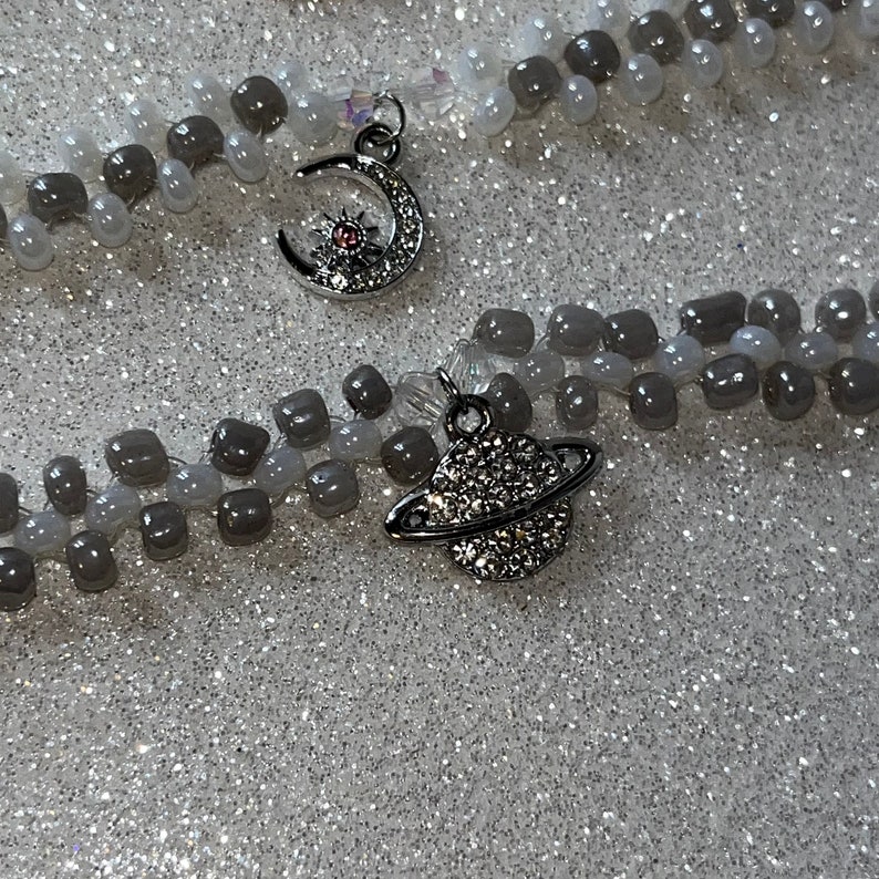 May include: Two beaded bracelets with silver charms. One charm is a crescent moon with a star and a pink gem. The other charm is a silver planet with a ring around it.