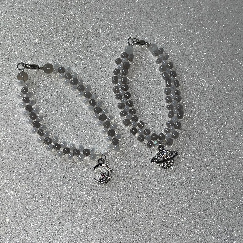 May include: Two beaded bracelets with silver charms. One charm is a crescent moon with a star, the other is a planet with a ring.