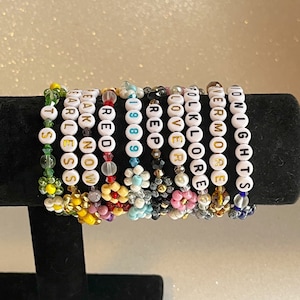 Pu&ograve; includere: Un set di 12 braccialetti colorati con perline bianche che compongono diverse parole, tra cui "Folklore", "Lover", "1989", "Reputation", "Red", "Speak Now", "Fearless", "Evermore", "Carlen", "Midnight" e "St. S", su una superficie nera.