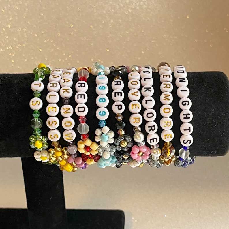 Pu&ograve; includere: Un set di 12 braccialetti colorati con perline bianche che compongono diverse parole, tra cui "Folklore", "Lover", "1989", "Reputation", "Red", "Speak Now", "Fearless", "Evermore", "Carlen", "Midnight" e "St. S", su una superficie nera.