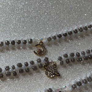May include: Two layered necklaces with gray and white beads. The top necklace has a gold crescent moon charm with a small sun in the center. The bottom necklace has a gold charm of a planet with a ring around it.