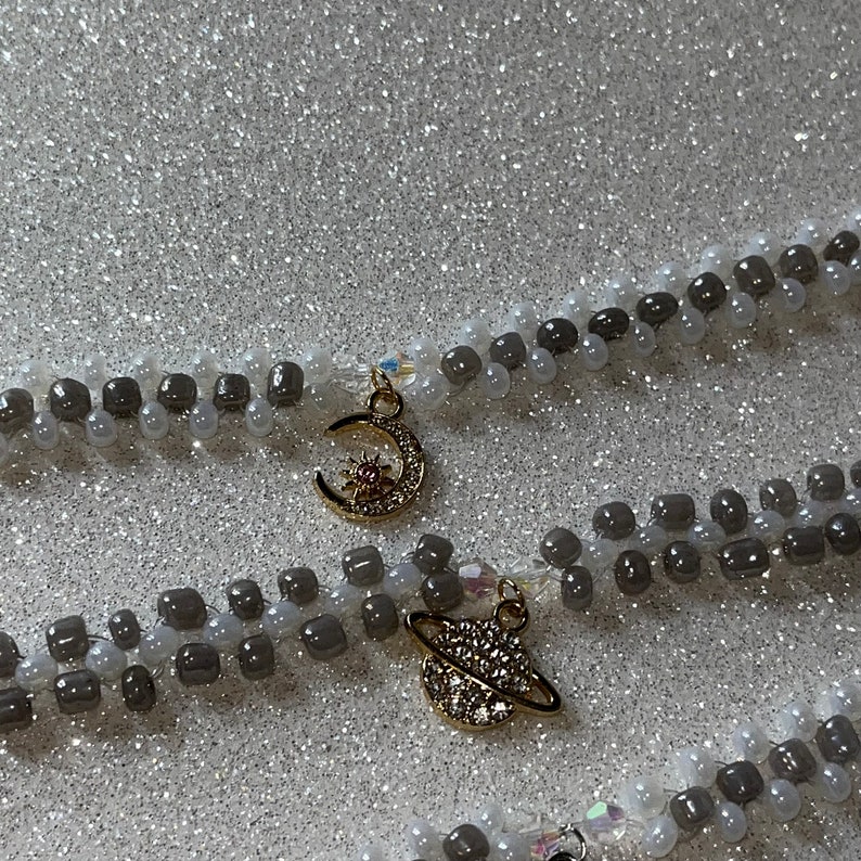 May include: Two layered necklaces with gray and white beads. The top necklace has a gold crescent moon charm with a small sun in the center. The bottom necklace has a gold charm of a planet with a ring around it.