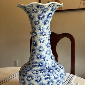 Puede incluir: Un jarrón grande de cerámica blanca con un patrón floral azul. El jarrón tiene una boca ancha y un cuello estrecho. Está sentado en una mesa blanca con un corredor a rayas gris y blanco.